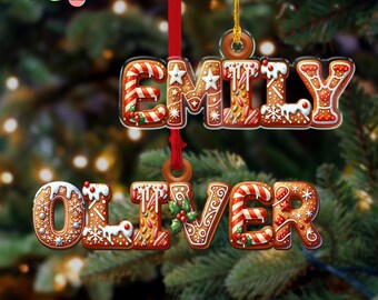 May include: Two gingerbread-style Christmas ornaments, one spelling "EMILY" and the other "OLIVER". The ornaments are decorated with white icing, candy canes, and sprinkles. They hang from red ribbons against a blurred Christmas tree background.