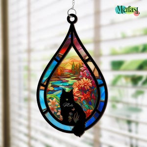 May include: A stained glass window hanging in a teardrop shape, featuring a black cat silhouette with the name "Mila" and a colorful landscape scene. The scene includes a river, flowers, and a sunset. The window is framed in black and has a silver chain for hanging.