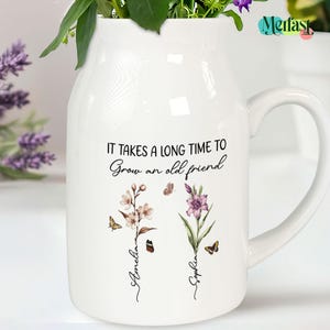 May include: White ceramic vase with a handle, featuring the quote "IT TAKES A LONG TIME TO Grow an old friend." Floral and butterfly illustrations are below the quote, with the names "Amelia" and "Sophia" in cursive. The vase is filled with greenery and purple flowers.