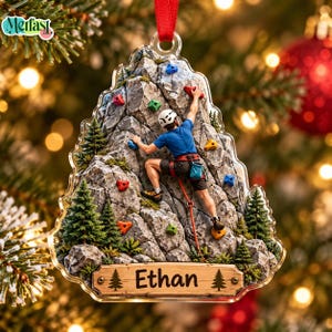 May include: A clear acrylic Christmas tree-shaped ornament featuring a rock climber in a blue shirt, black shorts, and white helmet scaling a rocky cliff with colorful climbing holds, green pine trees, and a wooden plaque engraved with the name Ethan.