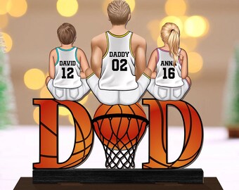 May include: A wooden basketball-themed family ornament. The ornament features a father and two children sitting on a basketball. The word "DAD" is displayed in large orange letters. The children's jerseys have the names "DAVID" and "ANN" with numbers 12 and 16.
