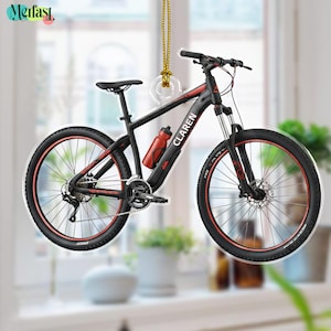 May include: A black and red mountain bike ornament with the word "CLAREN" on the frame. The bike has red accents on the wheels and frame, a red water bottle, and is hanging from a gold string.