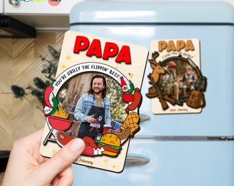 May include: A wooden magnet with the word "PAPA" in red letters. The magnet features a photo of a person wearing an apron and surrounded by cartoon images of grilling food. Text reads "You're Grilly the Flippin' Best" and "Johnny."