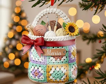 May include: A crochet basket-shaped Christmas ornament with a handle, filled with yarn balls, crochet hooks, and a sunflower. The basket is decorated with a red bow and the name "Maria" on a tag. The basket is made of colorful granny squares.