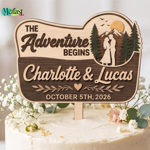 May include: A wooden cake topper featuring the text "THE Adventure BEGINS" and "Charlotte & Lucas" with the date "OCTOBER 5TH, 2026". The design includes a laser-cut scene of a couple standing before mountains, a sun, and pine trees, accented by a small heart.