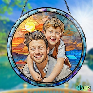 May include: Round stained glass window art featuring a father and son embracing, with a sunset and ocean backdrop. The artwork is framed with a black metal border and a chain for hanging. The image has a vibrant color palette.