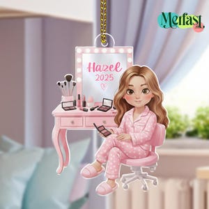 May include: A pink ornament featuring a cartoon illustration of a person in pink pajamas sitting at a vanity. The vanity has a mirror with the name "Hazel" and the year "2025". Makeup brushes, palettes, and other beauty products are on the vanity.