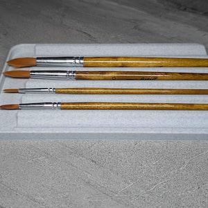 May include: Four paint brushes with wooden handles and silver ferrules are arranged in a light grey tray. The bristles are a light brown colour. The tray is rectangular with raised dividers to hold the brushes.