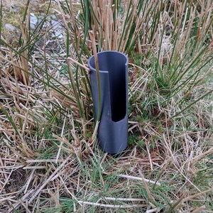 May include: A black plastic pipe with a slit in the side is partially buried in the ground. The pipe is surrounded by dry grass and green blades of grass.