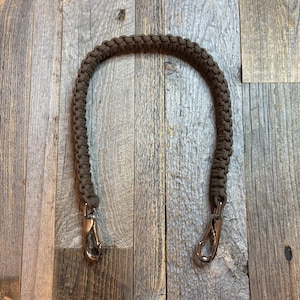 May include: A brown, braided paracord handle with silver-colored metal clasps. The handle is curved and appears sturdy, designed for attaching to a bag or other item. The background is a wooden surface.