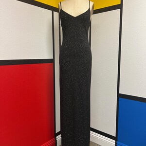 May include: A floor-length black slip dress featuring thin spaghetti straps and a V-neckline. The fabric is adorned with a subtle, repeating wavy pattern of tiny silver-toned dots that create a shimmering effect across the entire garment.
