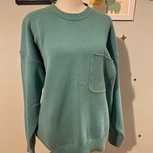 May include: A teal blue knitted sweater with a single pocket on the front. The sweater has a ribbed neckline and cuffs.