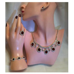 May include: A jewelry set consisting of a silver-toned necklace with oval red stones and clear crystals, a matching bracelet with oval red stones, a ring with a single oval red stone, and a single stud earring with an oval red stone.