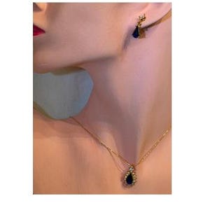 May include: A gold-tone necklace featuring a teardrop-shaped dark blue pendant surrounded by small clear stones, paired with a matching earring that has a similar teardrop blue stone and a clear stone accent on a gold-tone post setting.