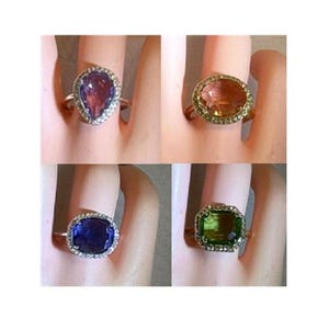 May include: A collection of four rings featuring large faceted center stones in pear, oval, cushion, and emerald cuts. Each ring is surrounded by a halo of small clear stones set on a thin metal band, showcasing purple, orange, blue, and green hues.