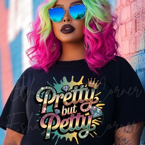 May include: Black t-shirt with the text "Pretty but Petty" in a colorful, jeweled design. The shirt features a crown and diamond graphic. The person in the image is wearing blue mirrored sunglasses and has pink and green hair.