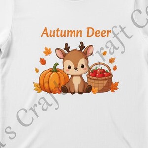 May include: White t-shirt featuring a cartoon deer, pumpkin, and basket of apples. The text "Autumn Deer" is above the image. Autumn leaves are scattered around the design. The shirt is a classic crew neck.