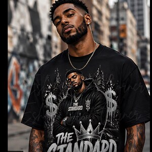 May include: Black t-shirt featuring a graphic of a man in a black jacket, a crown, and the words "THE STANDARD" in silver. The design includes dollar signs and a cityscape. The person in the image is wearing a silver chain.