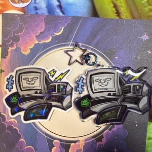 May include: Two computer-shaped keyrings with a smiling face on the screen, surrounded by lightning bolts and other graphics. The keyrings are silver and black with a holographic effect. A star-shaped charm and a blue ring are attached.