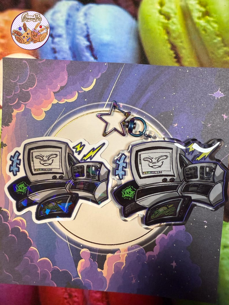 May include: Two computer-shaped keyrings with a smiling face on the screen, surrounded by lightning bolts and other graphics. The keyrings are silver and black with a holographic effect. A star-shaped charm and a blue ring are attached.