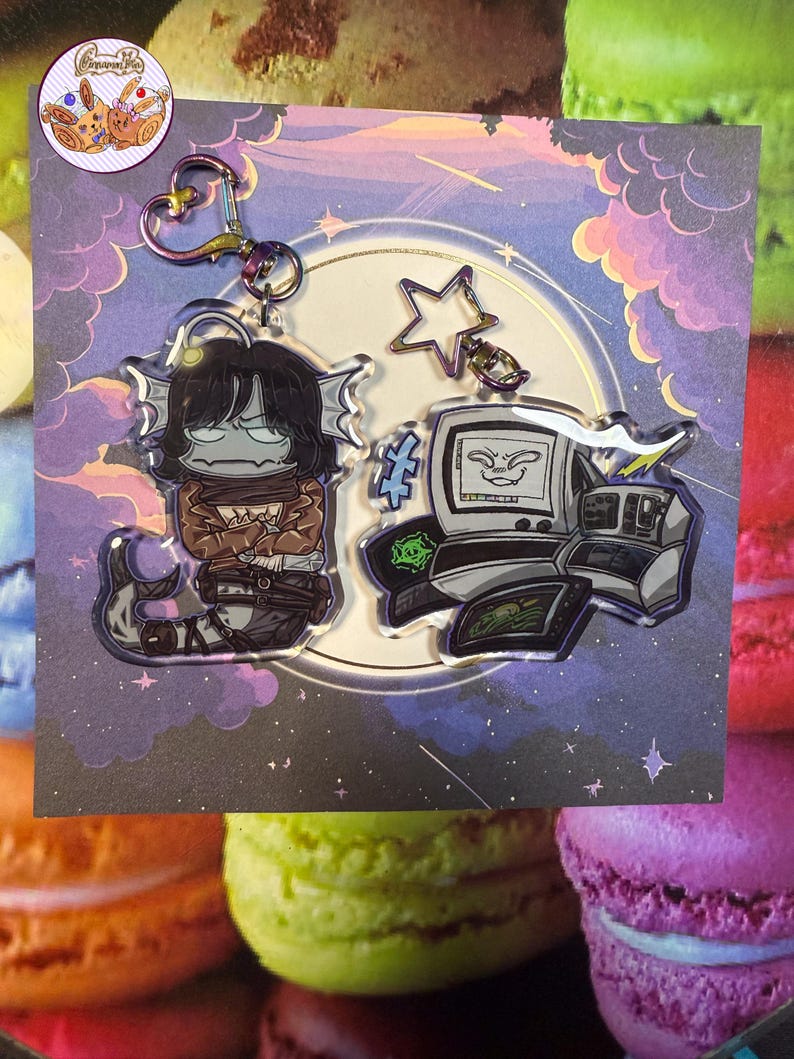 May include: Two acrylic keychains with cartoon characters. One shows a creature with dark hair and fish-like features, the other a retro computer with a face. Both are attached to a silver clasp and a star-shaped charm.