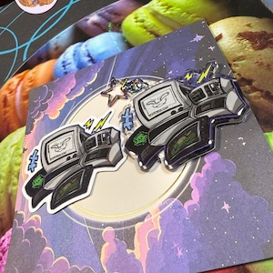 May include: Two enamel pins with a retro computer design, a smiling face on the screen, lightning bolts, and a star. The pins are set against a backdrop of a purple and blue night sky with clouds.