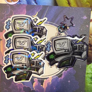 May include: A collection of stickers and a keychain showcasing a retro computer design with a smiling face on the screen. The stickers and keychain have a holographic effect, with lightning bolts and a star-shaped charm.