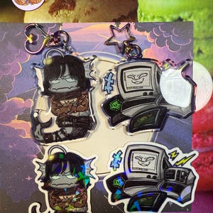 May include: Keyrings showcasing two designs: a fish-like creature and a retro computer. The fish-like character has black hair and a brown jacket. The computer design has a screen with a face. Both keyrings have iridescent accents and metal hardware.