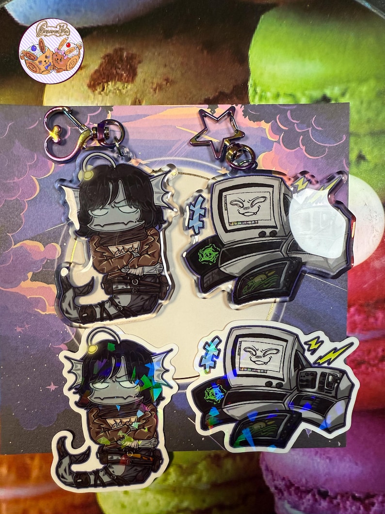 May include: Keyrings showcasing two designs: a fish-like creature and a retro computer. The fish-like character has black hair and a brown jacket. The computer design has a screen with a face. Both keyrings have iridescent accents and metal hardware.