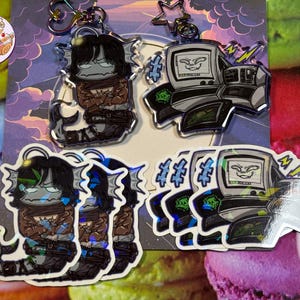 May include: A selection of stickers and keyrings with cartoon illustrations. The keyrings show a creature with a fish tail and a computer. The stickers depict the same characters. The colour scheme includes black, grey, brown, and blue.