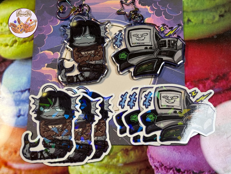 May include: A selection of stickers and keyrings with cartoon illustrations. The keyrings show a creature with a fish tail and a computer. The stickers depict the same characters. The colour scheme includes black, grey, brown, and blue.