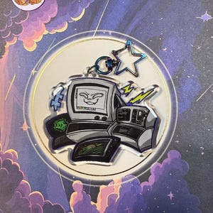 May include: A clear acrylic charm with a retro computer design, featuring a smiling face on the monitor. The charm includes a star-shaped charm and lightning bolt accents, attached to a silver keyring. The background has a celestial theme.