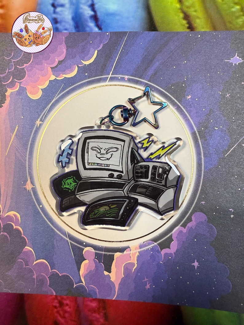May include: A clear acrylic charm with a retro computer design, featuring a smiling face on the monitor. The charm includes a star-shaped charm and lightning bolt accents, attached to a silver keyring. The background has a celestial theme.