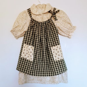 Könnte beinhalten: Ein Kleid im Vintage-Stil für Kinder. Das Oberteil ist cremefarben mit floralem Muster, der Rock ist schwarz-grün kariert. Es hat kurze, gepuffte Ärmel, einen Bubikragen und zwei kleine Taschen mit floralen Akzenten. Auf einem weißen Kleiderbügel.