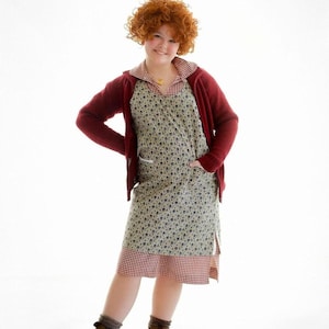 Könnte beinhalten: Eine Frau trägt eine rote Strickjacke über einem Kleid mit floralem Muster und einem rot-weiß karierten Saum. Das Kleid hat Taschen und wird mit braunen Stiefeln und grauen Socken kombiniert. Die Frau hat rotes lockiges Haar und eine goldene Halskette.