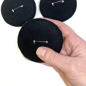 May include: Three black felt circles with a silver safety pin attached to each. The circles are approximately 3 inches in diameter.