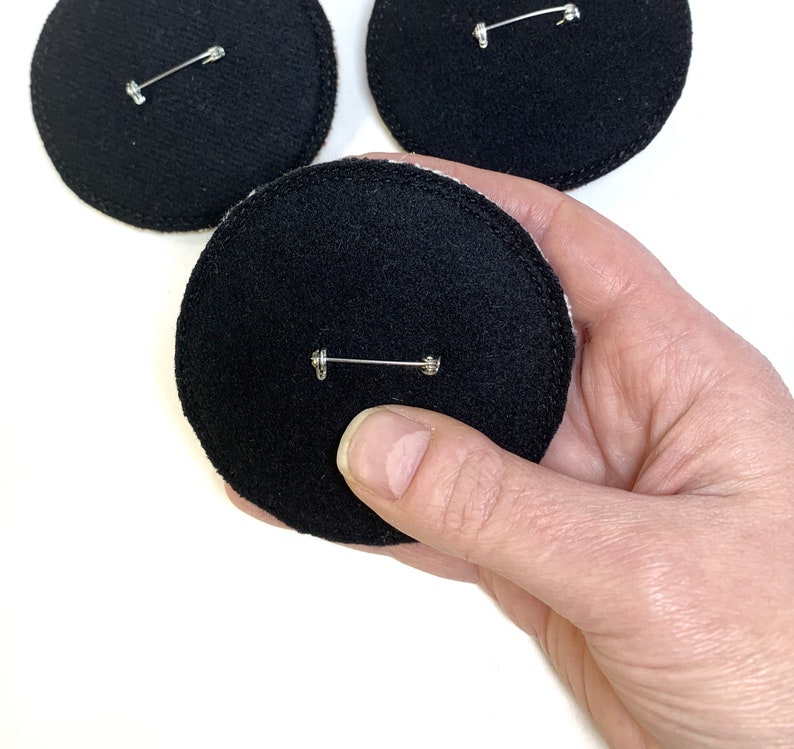 May include: Three black felt circles with a silver safety pin attached to each. The circles are approximately 3 inches in diameter.