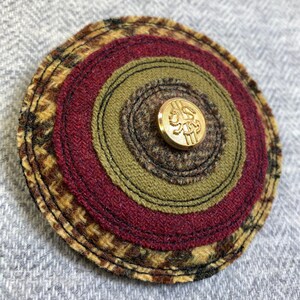 May include: A circular fabric wall hanging with concentric circles of brown, green, and red fabric. A gold button with a lion design is in the center.