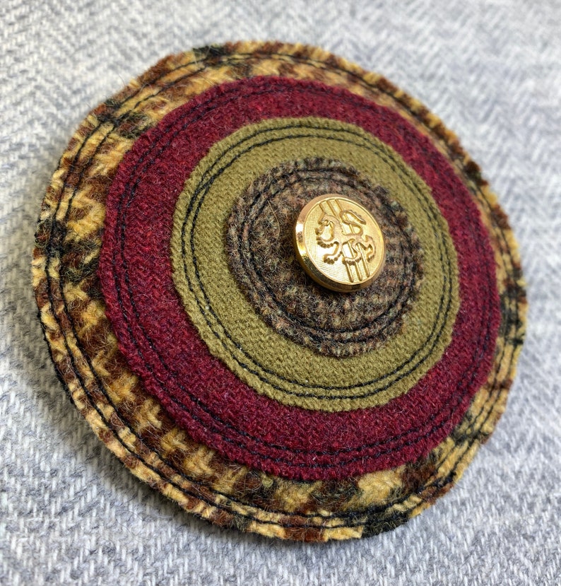 May include: A circular fabric wall hanging with concentric circles of brown, green, and red fabric. A gold button with a lion design is in the center.