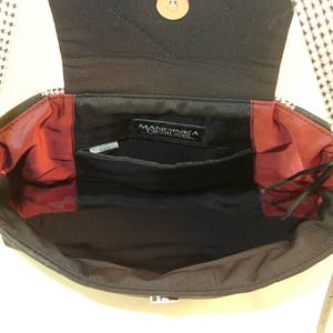 May include: Black shoulder bag with a flap closure, featuring a black and white patterned strap. The interior is black with red accents and multiple pockets. The bag has a metal clasp and a gold-colored button.
