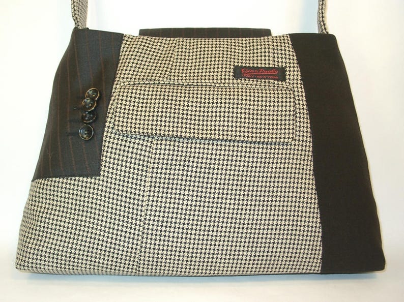 May include: A rectangular tote bag made from repurposed suit materials. The bag features a houndstooth pattern, pinstriped fabric, and black accents. It has a pocket with a label and buttons on the side. The bag has a shoulder strap.