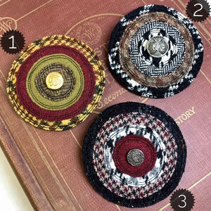 May include: Three round fabric brooches with different patterns and colors. Each brooch has a button in the center. The brooches are on a brown book with the word "Second" visible.