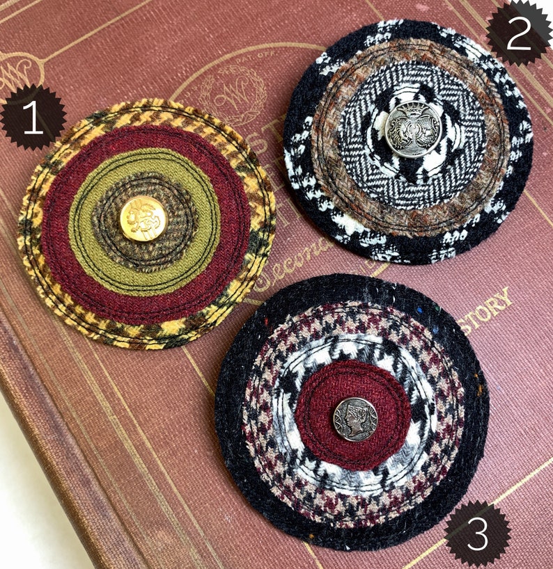 May include: Three round fabric brooches with different patterns and colors. Each brooch has a button in the center. The brooches are on a brown book with the word "Second" visible.