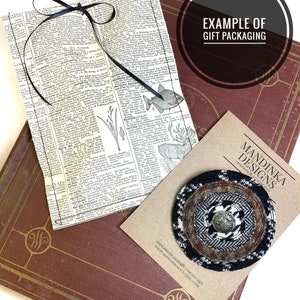 May include: A brown and black patterned fabric pin with a silver button in the center. The pin is on a brown card with the text "MANDINKA DESIGNS" and "HANDMADE & COZY" in white. The card also includes the website address "MANDINKADESIGNS.COMCAST.NET" and "WWW.MANDINKADESIGNS.COM".