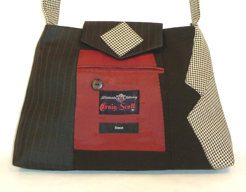 May include: A unique shoulder bag crafted from repurposed suit fabrics. The bag features a black and brown pinstripe pattern, a red pocket with a button, and a houndstooth flap. The label reads "Craig Scott" and "Stretch".