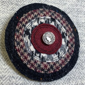 May include: A round, multi-layered fabric pin with a black, red, and white pattern. The center of the pin features a silver button with a raised design of a woman's profile.