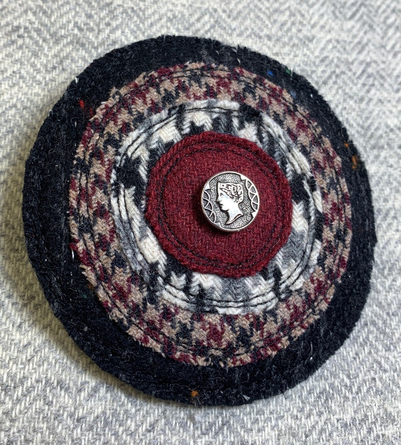 May include: A round, multi-layered fabric pin with a black, red, and white pattern. The center of the pin features a silver button with a raised design of a woman's profile.