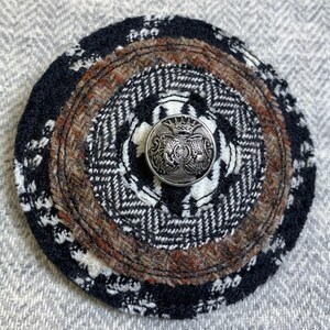 May include: A black and brown circular wool patch with a silver button in the center. The button has a crown and a design of three stylized figures.