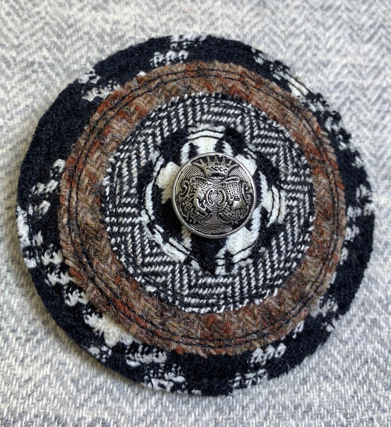 May include: A black and brown circular wool patch with a silver button in the center. The button has a crown and a design of three stylized figures.