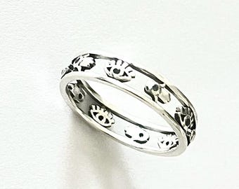 May include: A silver ring with an eye pattern. The ring is made of a shiny metal and features a repeating design of stylized eyes around the band. The ring is shown on a white background.